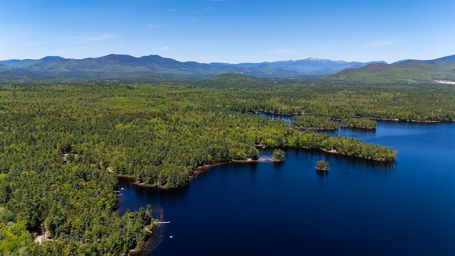 This high-angle aerial perspective captures a serene lakefront landscape surrounded by dense, lush green forests. The deep blue water reflects the clear sky, with small islands and a rugged shoreline adding natural character to the scene. In the distance, rolling mountain ranges provide a majestic backdrop, emphasizing the property's secluded and scenic location.