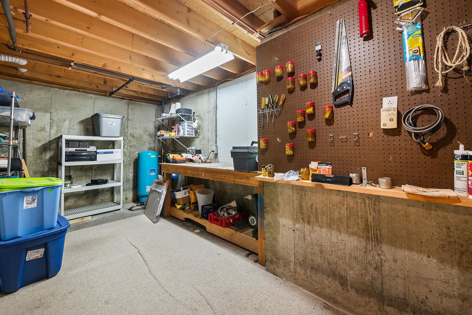 This unfinished basement space features a functional workshop area with a long wooden workbench and a large pegboard wall organized with various tools and storage jars. The room includes exposed wooden ceiling joists, concrete foundation walls, and a carpeted floor, providing a practical utility space. A white metal shelving unit and blue storage bins offer additional organization, while a bright overhead light fixture illuminates the workspace.
