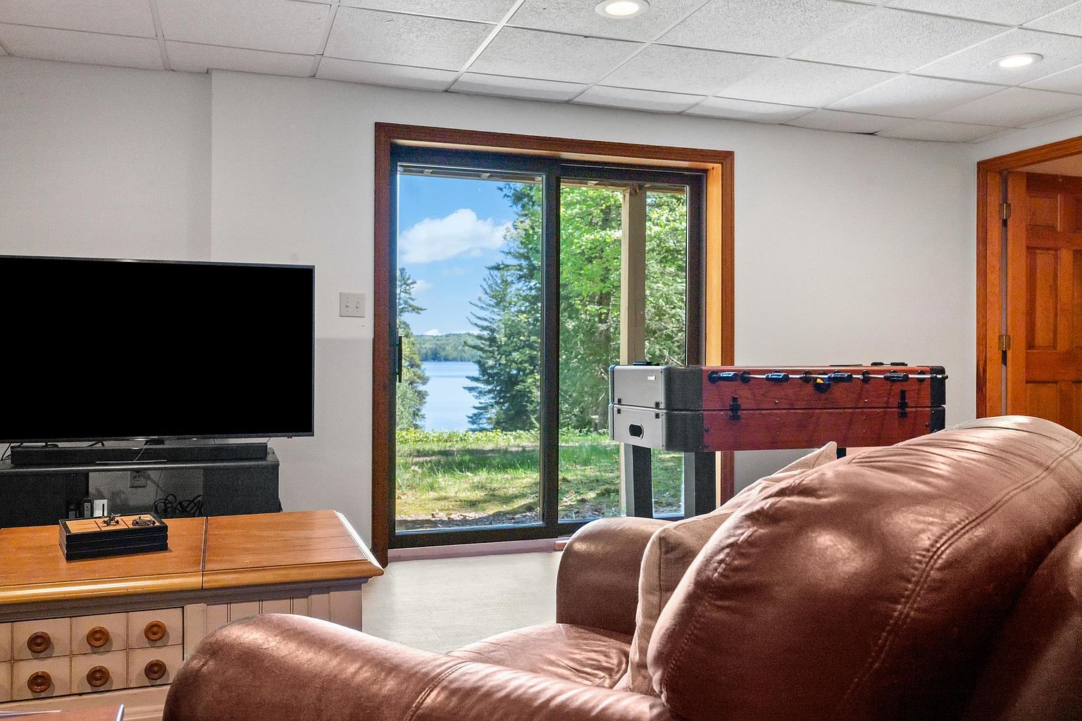 This interior view of a basement living area features a comfortable brown leather sofa in the foreground and a television set on a media console. A sliding glass door provides a clear view of a scenic lake and surrounding trees, bringing natural light into the space. To the right, a foosball table adds a recreational element to the room, which is finished with a drop ceiling and neutral walls.