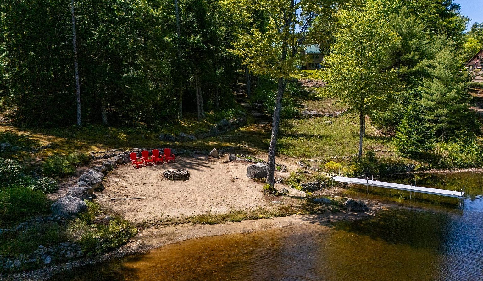 This aerial perspective captures a serene lakeside property featuring a sandy beach area complete with a stone fire pit and four bright red Adirondack chairs. A long, narrow dock extends from the shoreline into the clear water, while a gentle slope leads up through a wooded landscape toward a secluded house nestled among the trees. The scene evokes a peaceful, private retreat atmosphere, highlighting the natural beauty and recreational potential of the waterfront setting.