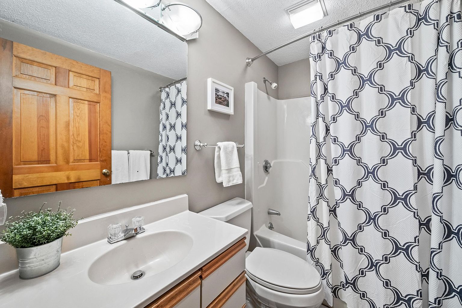 This compact, functional bathroom features a light-toned vanity with a white countertop and a single sink, complemented by a large wall-mounted mirror. A shower-tub combination is enclosed by a patterned, geometric-print shower curtain, and the walls are painted in a neutral gray tone. The space is well-lit by a vanity light fixture and a ceiling light, creating a clean and tidy atmosphere.