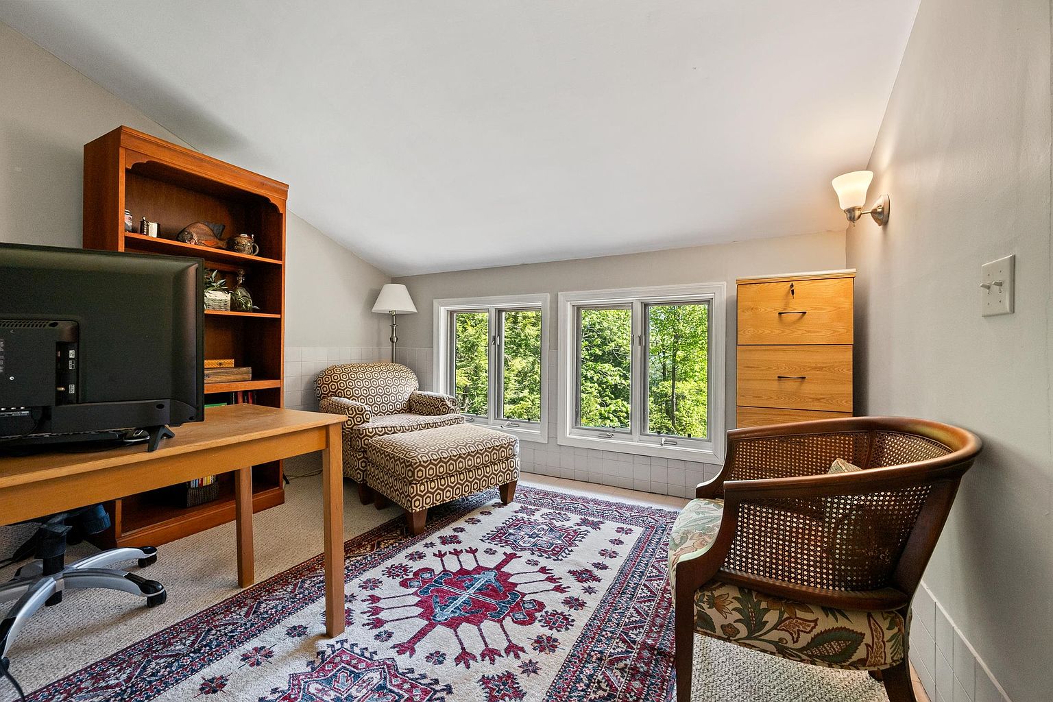 This cozy home office or study features a desk with a computer monitor, a tall wooden bookshelf, and a patterned chaise lounge positioned near two windows that offer a view of the trees outside. A vintage-style area rug anchors the space, which also includes a wooden chair with a patterned cushion and a light wood filing cabinet. The room has a sloped ceiling and neutral walls, creating a quiet and functional workspace.