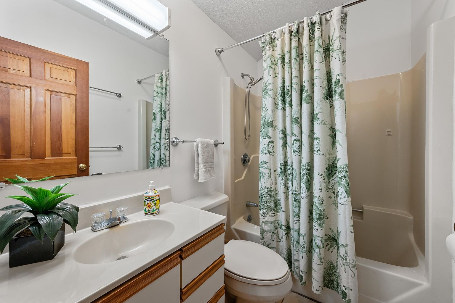 This functional bathroom features a single-sink vanity with light wood-toned trim and a large wall-mounted mirror. A shower-tub combination is enclosed by a patterned green and white shower curtain, complemented by a matching soap dispenser on the counter. The space is well-lit by a long vanity light fixture, creating a clean and practical atmosphere.