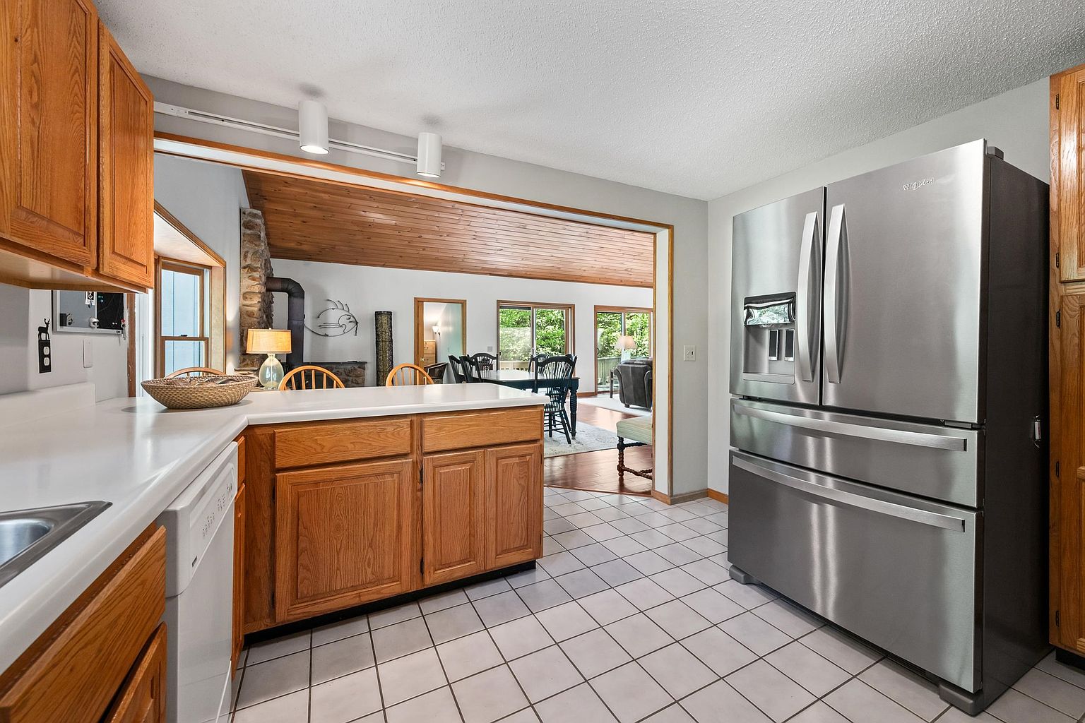 This kitchen features warm-toned wood cabinetry paired with white countertops and a white tiled floor. A stainless steel refrigerator stands prominently on the right, while an open-concept layout provides a clear view into the adjacent dining and living areas. The space is bright and functional, with a neutral color palette that creates a welcoming atmosphere.