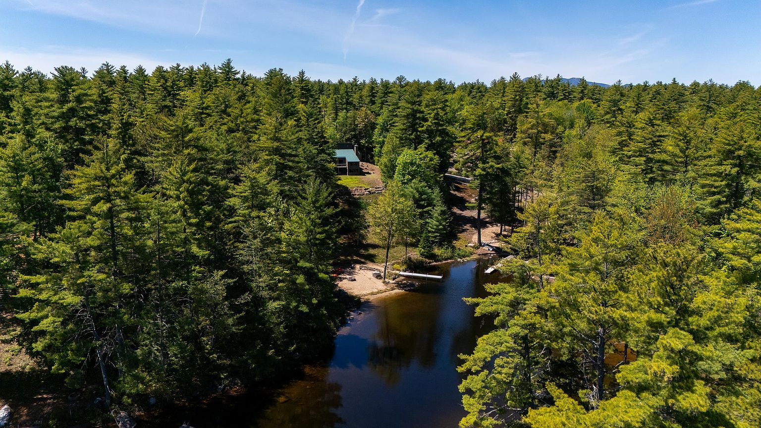 This high-angle aerial perspective captures a secluded cabin nestled within a dense, lush forest overlooking a tranquil lake. The property features a private sandy beach area and a wooden dock extending into the water, emphasizing a peaceful, nature-focused retreat. The surrounding landscape is dominated by mature evergreen trees, highlighting the home's private and serene setting.