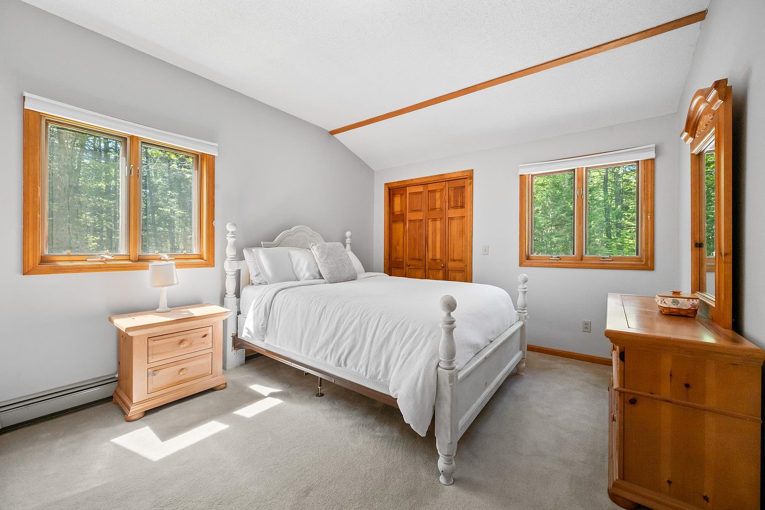 This bright bedroom features a white bed frame centered between two windows that look out onto a wooded area. The room is furnished with a light wood nightstand and a matching dresser with an attached mirror, complemented by neutral carpeting and light-colored walls. A wooden closet door adds a warm, traditional touch to the space, which is illuminated by natural light.