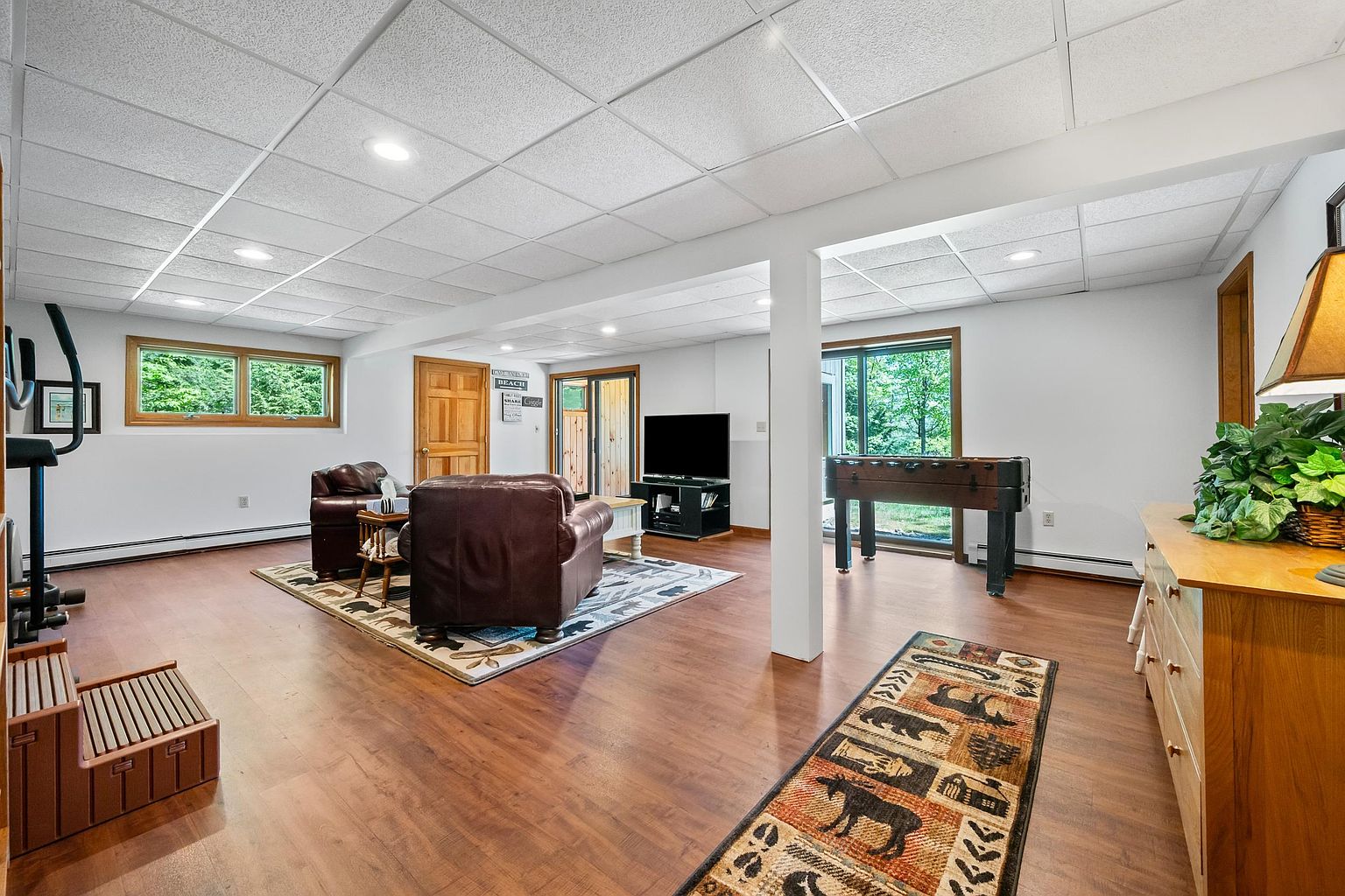 This spacious, finished basement features wood-look flooring and a drop ceiling with recessed lighting, creating a versatile living area. The room is furnished with leather seating around a television, a foosball table near the sliding glass doors, and a wooden dresser topped with a plant. The layout is open and airy, with large windows and glass doors providing natural light and views of the surrounding greenery.
