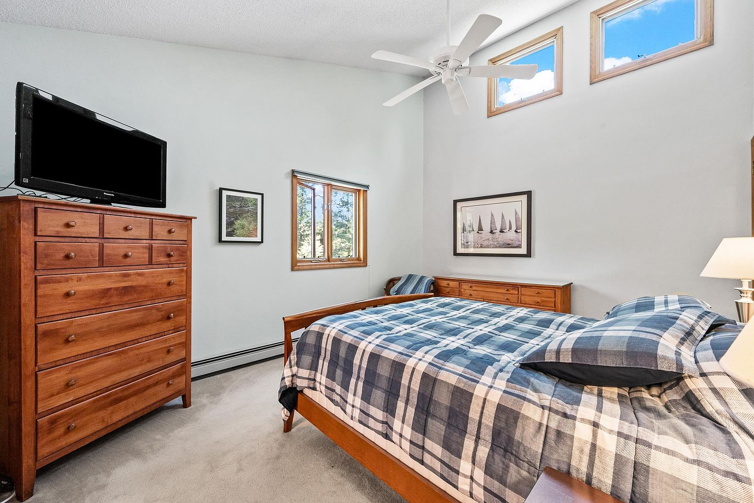 This bedroom features a vaulted ceiling with a white ceiling fan and two high-placed windows that allow for natural light. The room is furnished with a wooden dresser topped with a television, a matching bed frame with a blue and white plaid comforter, and a low-profile dresser against the back wall. A framed picture of sailboats hangs above the rear dresser, and the space is finished with neutral carpeting and light-colored walls.