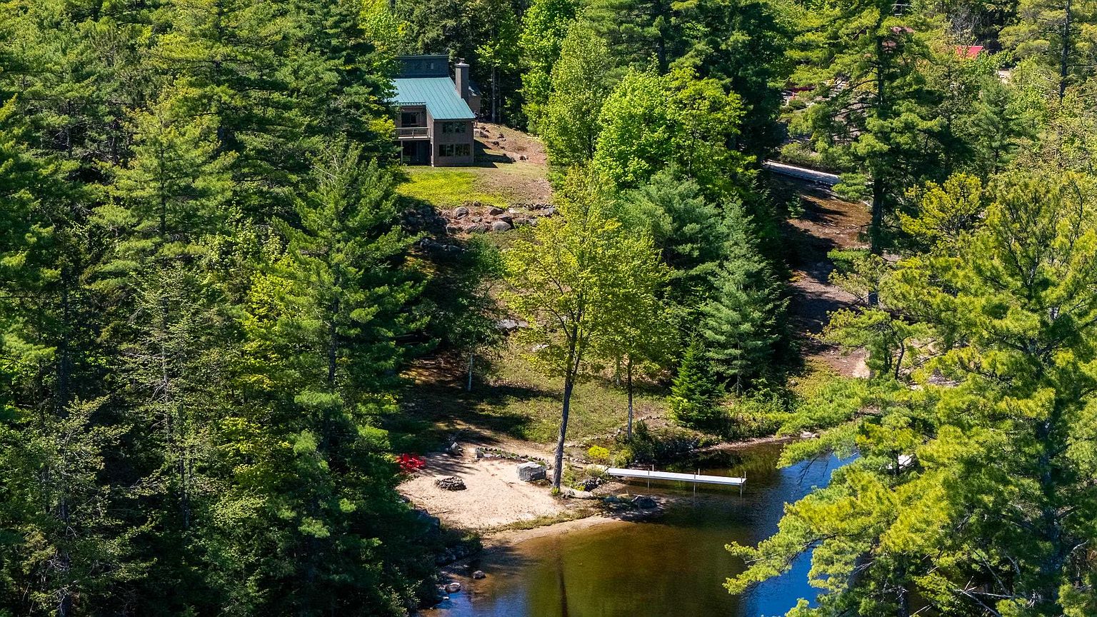 This aerial perspective captures a secluded, multi-level home nestled within a dense, lush forest overlooking a private waterfront. The property features a sandy beach area, a long wooden dock extending into the water, and a natural landscape that seamlessly integrates the residence into its wooded surroundings. The elevated vantage point highlights the home's peaceful, private setting and its direct access to the lake.