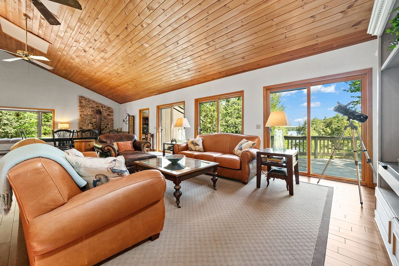 This spacious living room features a vaulted wood-paneled ceiling and a large stone-clad chimney breast, creating a warm, rustic atmosphere. The room is furnished with comfortable leather seating, a central coffee table, and a telescope positioned near large sliding glass doors that offer a scenic view of the outdoors. The space is bright and inviting, with a neutral area rug grounding the seating area and light wood flooring throughout.