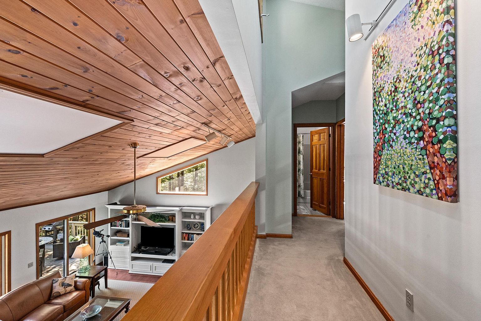 This perspective captures a carpeted upper-level hallway that overlooks a living area below, defined by a dramatic vaulted ceiling with exposed wood planking. The space features a wooden railing, a large abstract art piece on the wall, and a view into a lower-level living room equipped with built-in shelving and a ceiling fan. The architectural design creates an open, airy atmosphere that connects the different levels of the home.