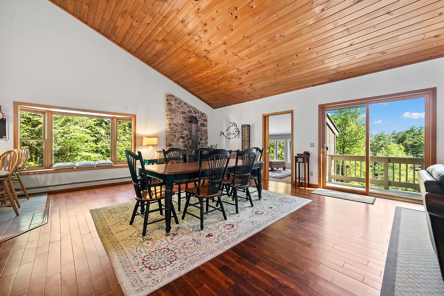 This dining room features a vaulted ceiling with warm wood paneling and a prominent stone-clad chimney breast that serves as a focal point. A large wooden dining table with spindle-back chairs sits atop a patterned area rug, while a wide window seat and sliding glass doors provide ample natural light and views of the surrounding trees. The space is finished with rich-toned wood flooring, creating a cozy and inviting atmosphere.