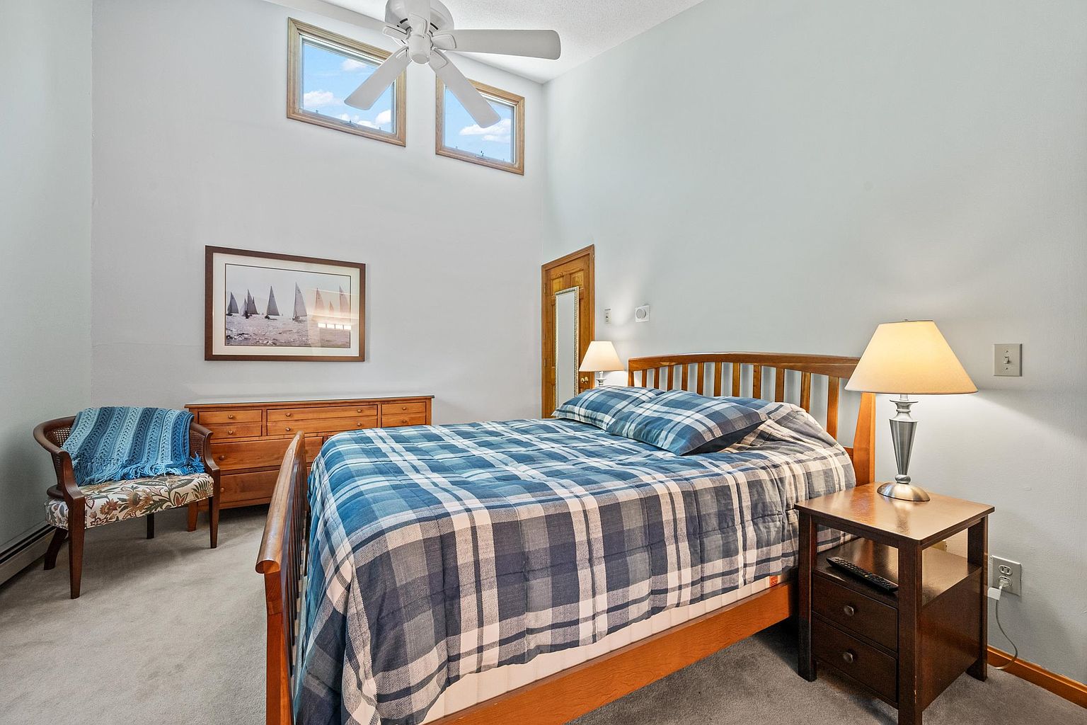 This bedroom features a wooden bed frame dressed in a blue and white plaid comforter, complemented by a matching nightstand with a lamp. To the left, a wooden dresser sits beneath a framed sailboat print, accompanied by a patterned armchair draped with a blue throw blanket. The room is brightened by two high-placed windows and a ceiling fan, creating a cozy and inviting atmosphere.