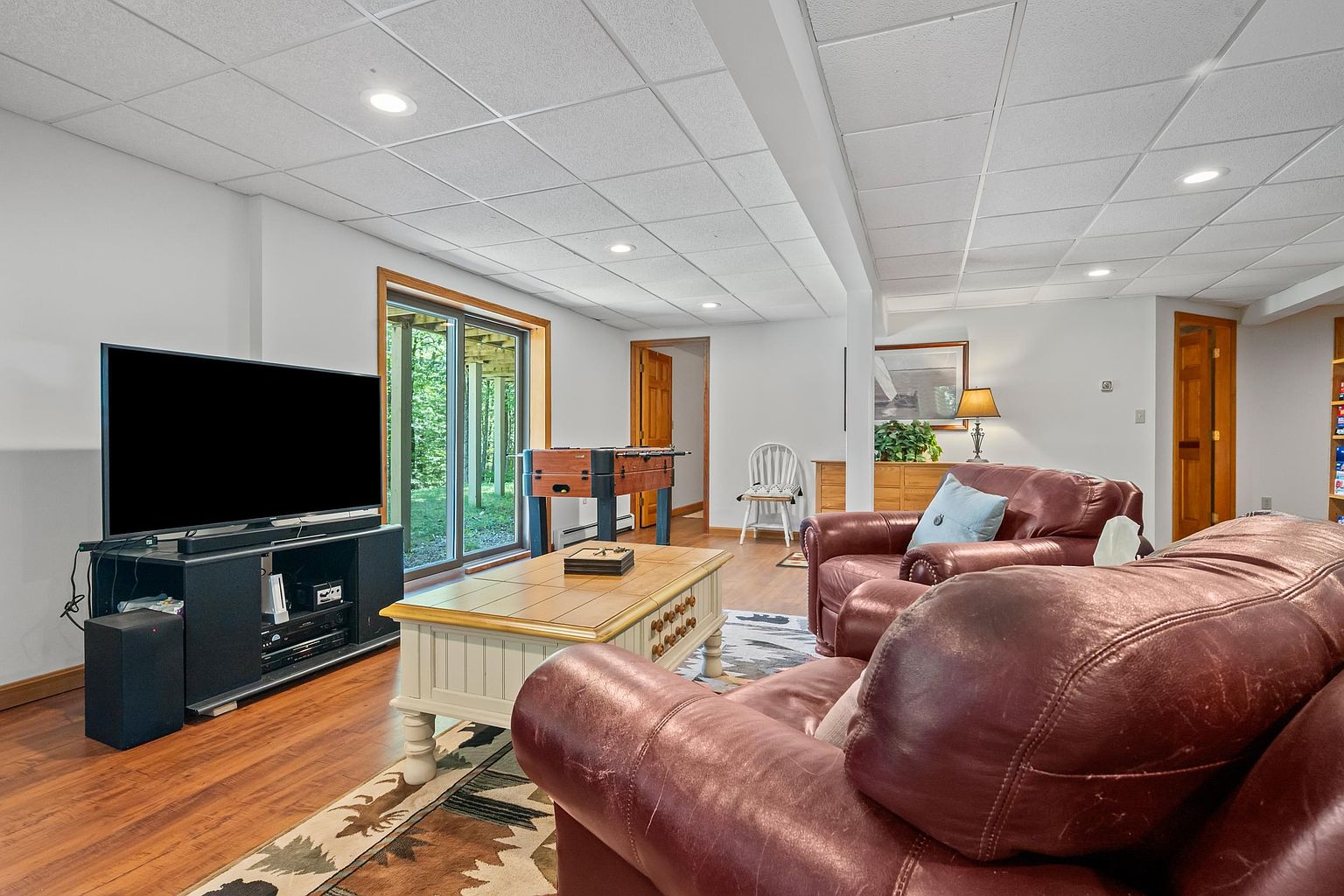 This spacious finished basement features a comfortable seating area with dark leather furniture arranged around a coffee table and a large television. The room includes a foosball table near the sliding glass doors that provide a view of the wooded backyard, and a white chair sits in the corner near a doorway. The space is finished with a drop ceiling, recessed lighting, and warm-toned wood flooring, creating a cozy and functional recreational area.