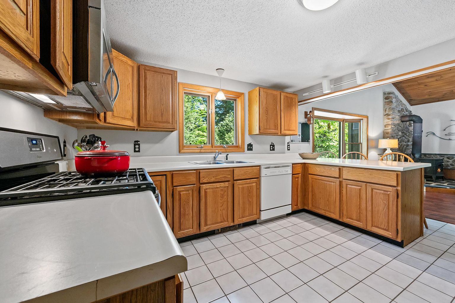 This kitchen features light wood cabinetry, white countertops, and a white tiled floor, creating a bright and functional workspace. A red pot sits on the gas range in the foreground, while a window above the sink provides natural light and a view of the outdoors. The space opens into an adjacent living area, which showcases a stone-clad chimney breast and a wood-burning stove, adding a rustic touch to the home's interior.