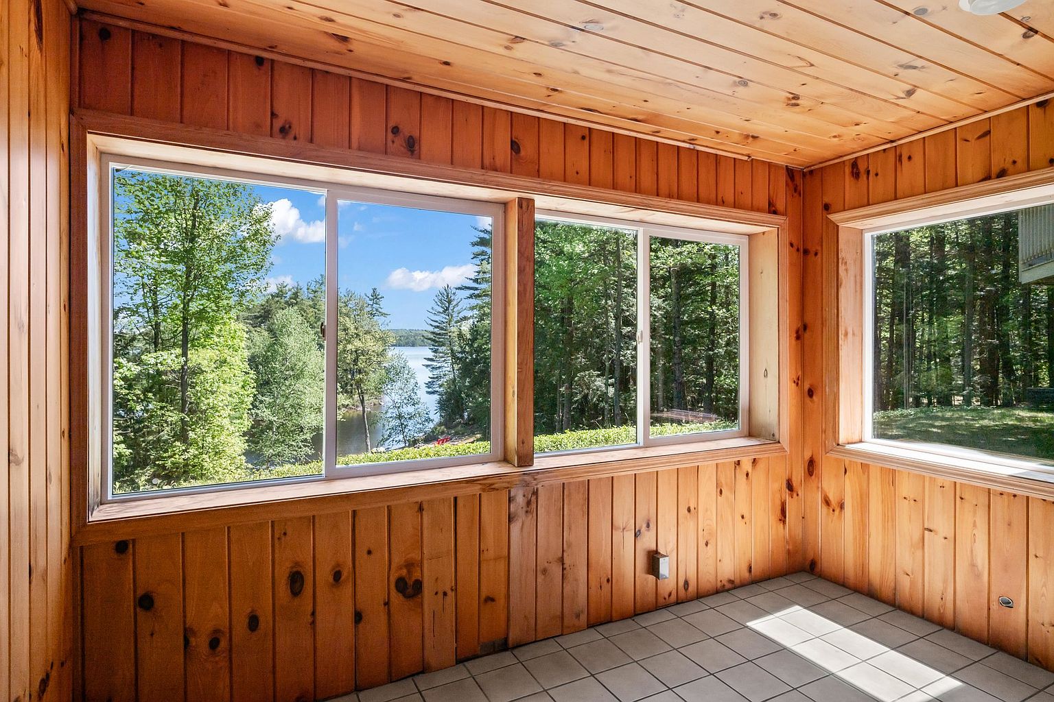 This sunroom features warm, light-toned wood paneling on the walls and ceiling, creating a cozy and rustic atmosphere. Large windows provide expansive, scenic views of the surrounding forest and a nearby lake, filling the space with natural light. The room is finished with neutral-toned square floor tiles, offering a clean and functional space to enjoy the outdoor scenery.