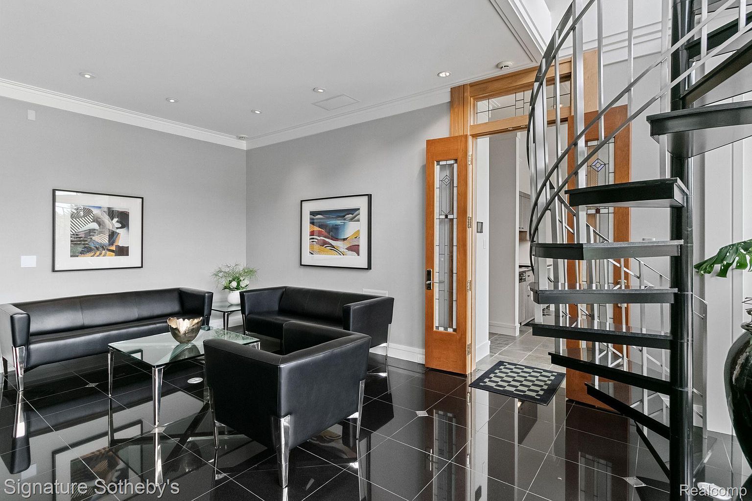 This is an interior shot of a modern living room featuring black leather sofas and chairs arranged around a glass coffee table. The room has a sleek, contemporary design with black tile flooring and artwork on the walls. A spiral staircase is visible in the background, adding an architectural element to the space.