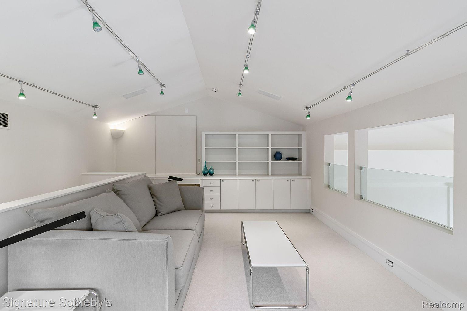 This is an interior shot of a modern living room featuring a light gray sofa, a white coffee table with a chrome frame, and built-in white shelving and cabinets. The room has a minimalist aesthetic with white walls and carpet, track lighting, and large windows with glass railings, creating a bright and airy atmosphere. The perspective is from a medium distance, showcasing the layout and key furniture pieces.