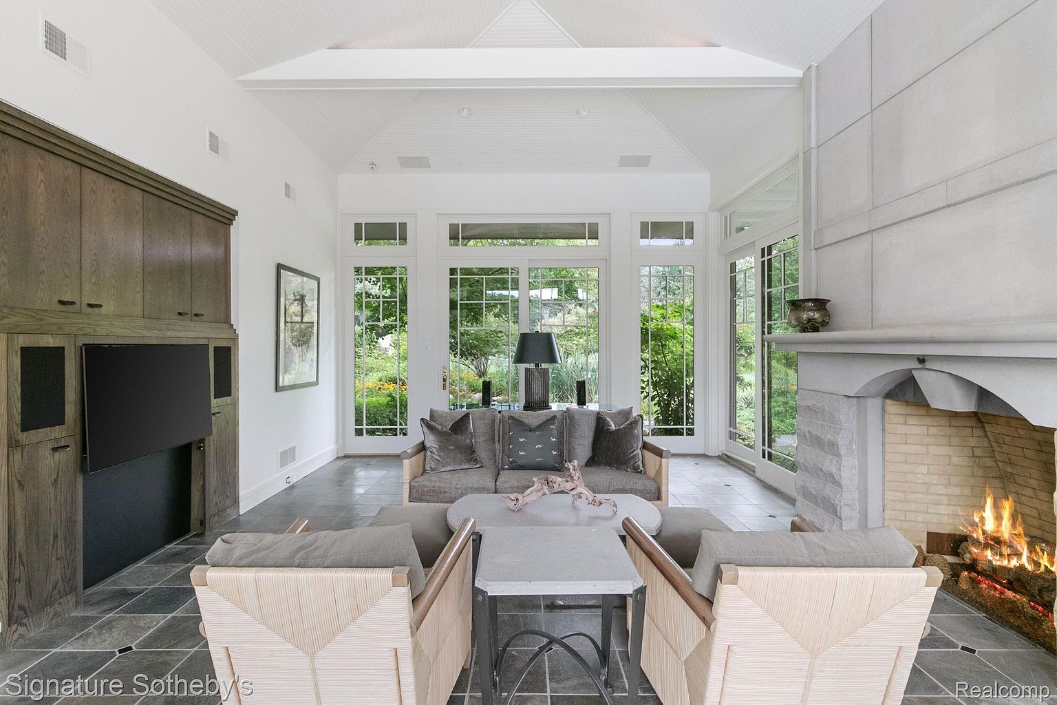 This is an interior shot of a sun-drenched living room featuring a fireplace, a built-in entertainment center, and large windows offering views of the garden. The room is furnished with a neutral-toned sofa, chairs, and a coffee table, creating a cozy and inviting atmosphere. The architectural details, such as the vaulted ceiling and stone fireplace, add to the room's charm and elegance.