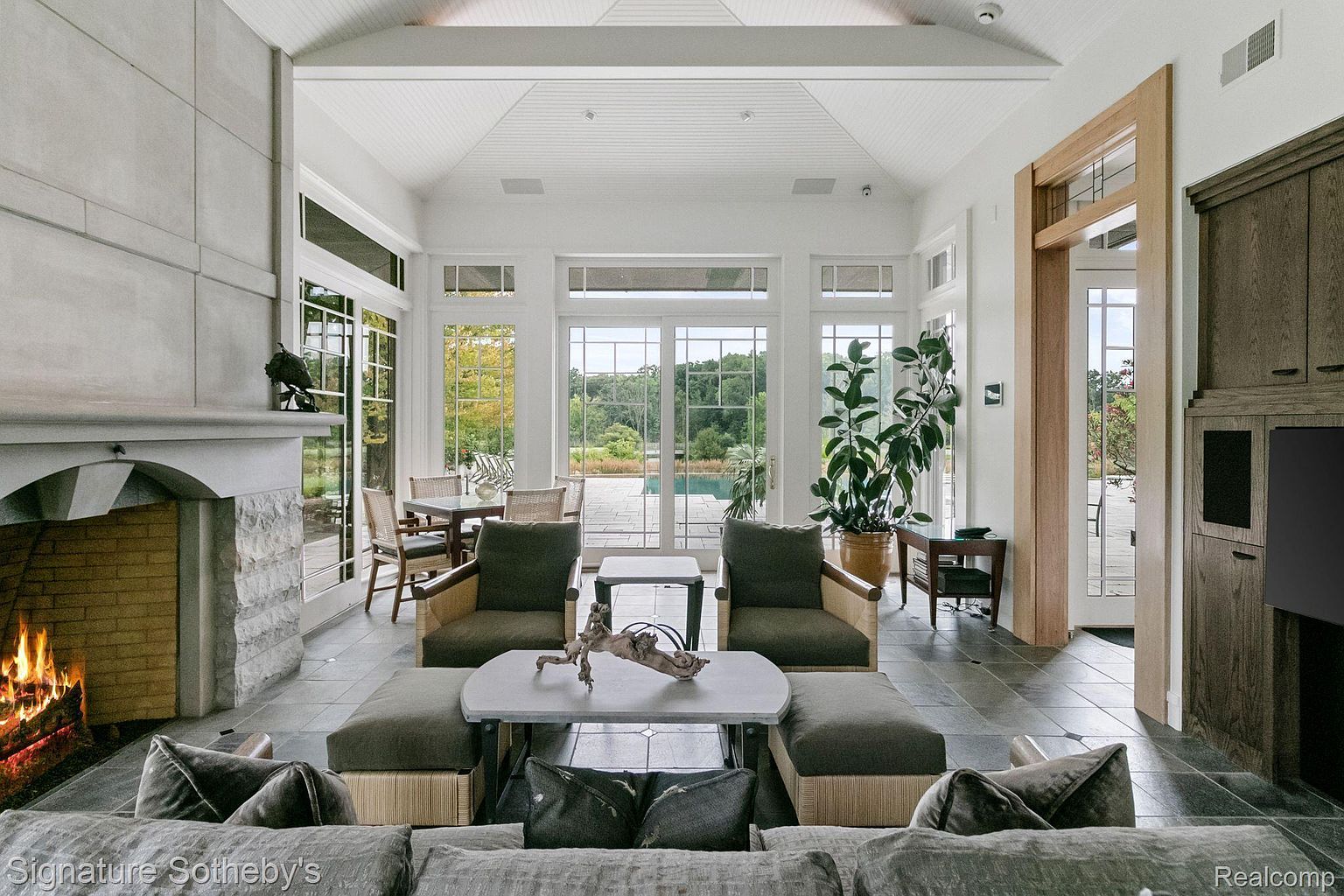This is an interior shot of a luxurious living room featuring a stone fireplace with a burning fire, large windows overlooking a lush outdoor area with a pool, and comfortable seating arrangements. The room has a high, white ceiling and a neutral color palette, creating a bright and airy atmosphere. The overall impression is one of elegance and comfort, perfect for relaxation and entertaining.