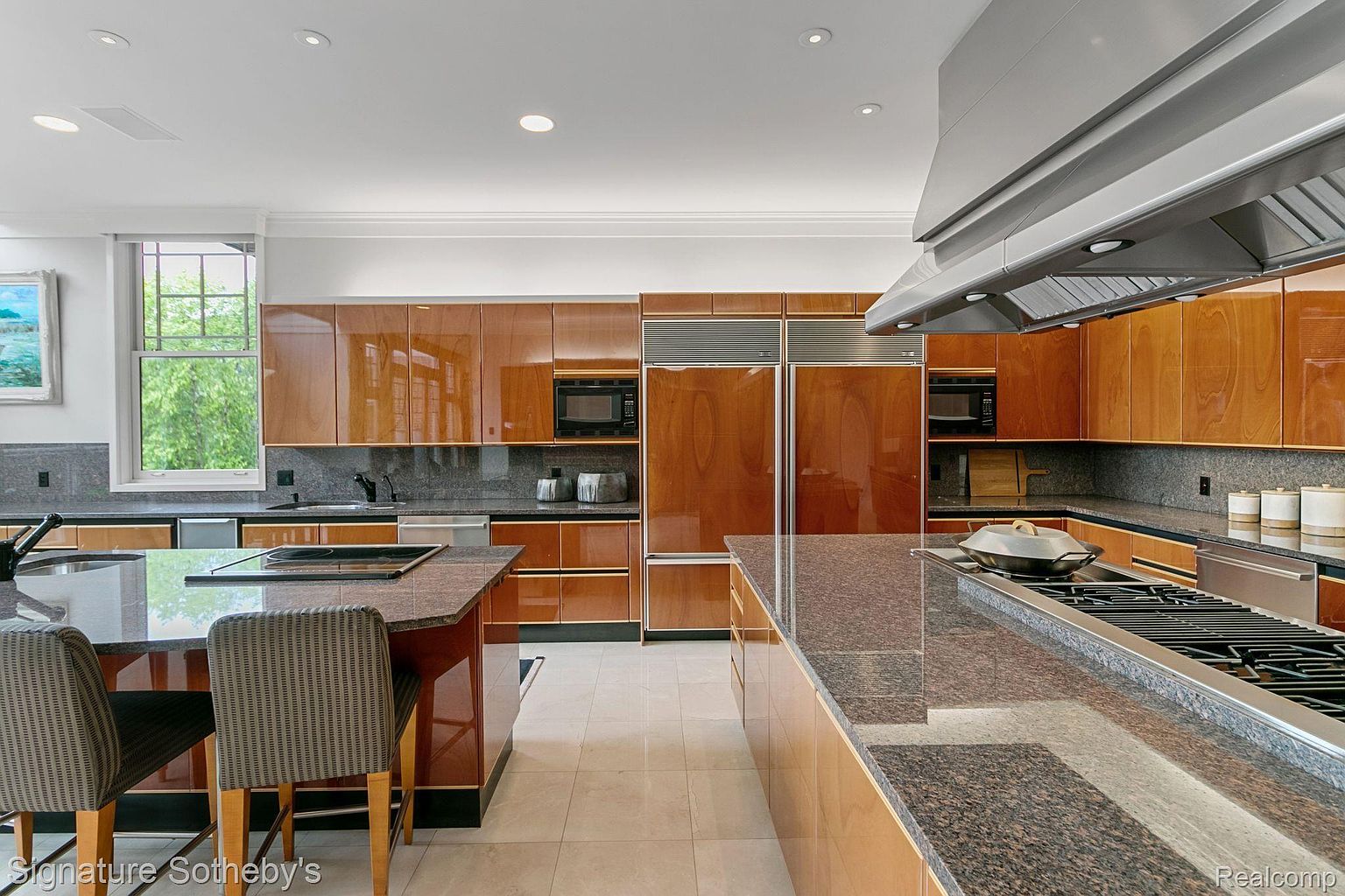 This is a modern kitchen with sleek, glossy wood cabinetry and dark granite countertops. Stainless steel appliances, including a large range and double refrigerators, are integrated into the design. The kitchen features a large island with seating and a cooktop, and the overall impression is one of luxury and functionality.