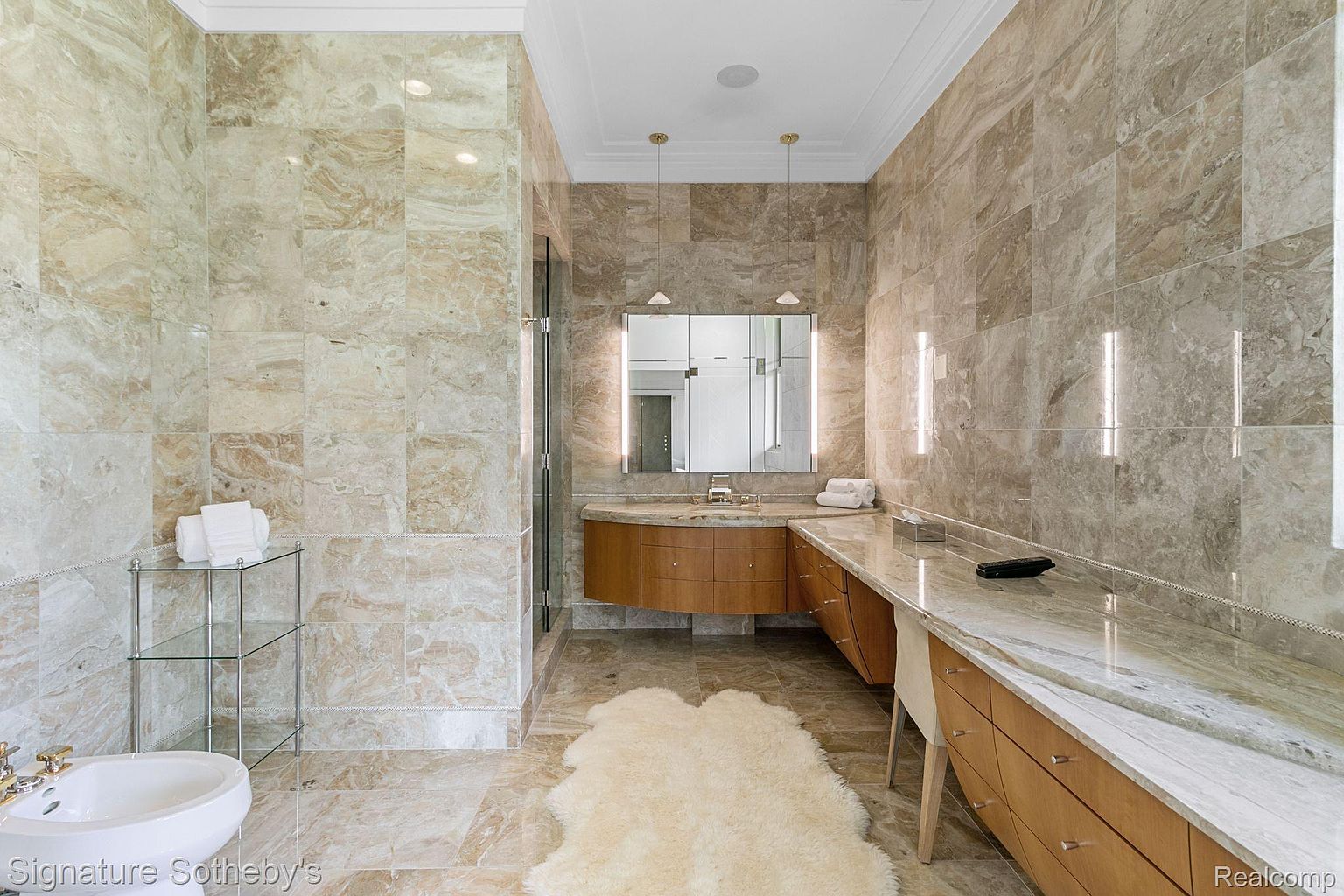 This is a luxurious primary bathroom featuring extensive use of beige marble on the walls and floors, creating a seamless and elegant look. The bathroom includes a vanity with wooden cabinets and a marble countertop, a large mirror with integrated lighting, and a bidet. A plush rug adds a touch of comfort to the space.