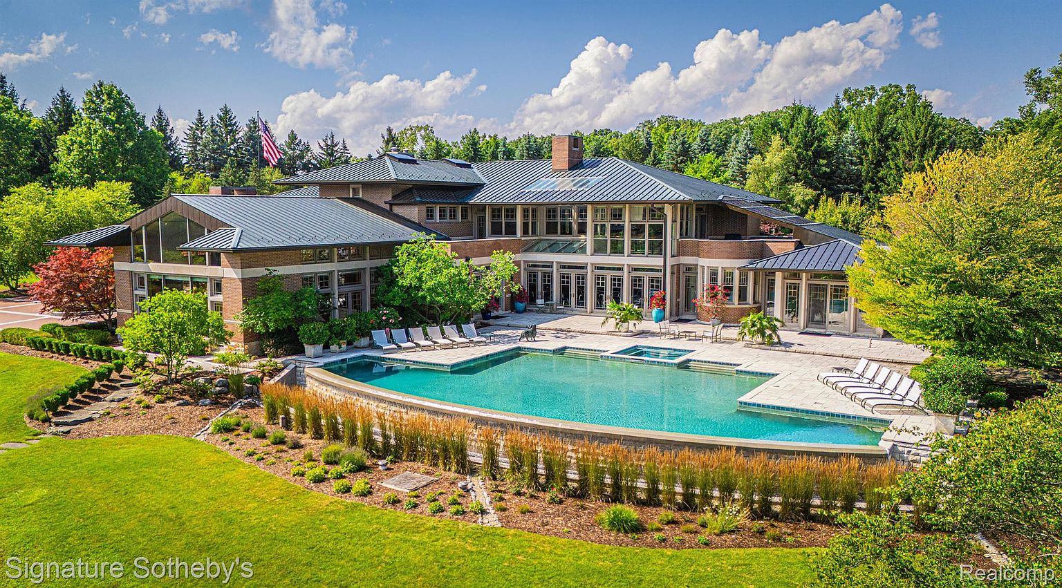 This is an aerial view of a luxurious home featuring a large, curved swimming pool and spa. The pool is surrounded by a stone patio with lounge chairs, and the house has a modern architectural style with a mix of brick and glass. Lush landscaping and mature trees provide privacy and enhance the property's appeal.