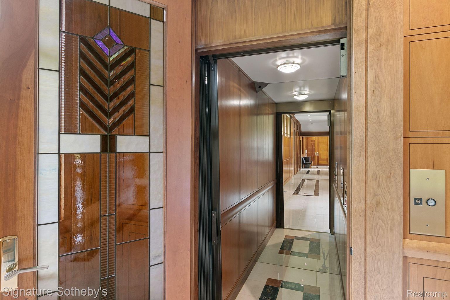 This interior shot showcases a hallway with rich wood paneling and a decorative stained glass window. An elevator is visible, suggesting luxury and convenience. The hallway leads to a brightly lit area with tiled flooring, creating a sense of depth and elegance.
