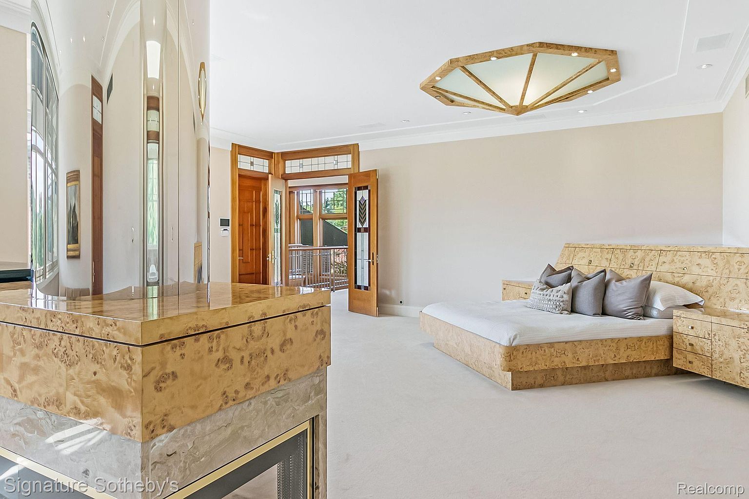 This primary bedroom features a unique, light-colored wood finish on the bed frame, headboard, and nightstands, creating a cohesive and luxurious aesthetic. The room is spacious with a neutral color palette, complemented by a striking geometric ceiling light fixture and an adjacent balcony area accessible through wooden doors. The overall impression is one of sophisticated comfort and distinctive design.