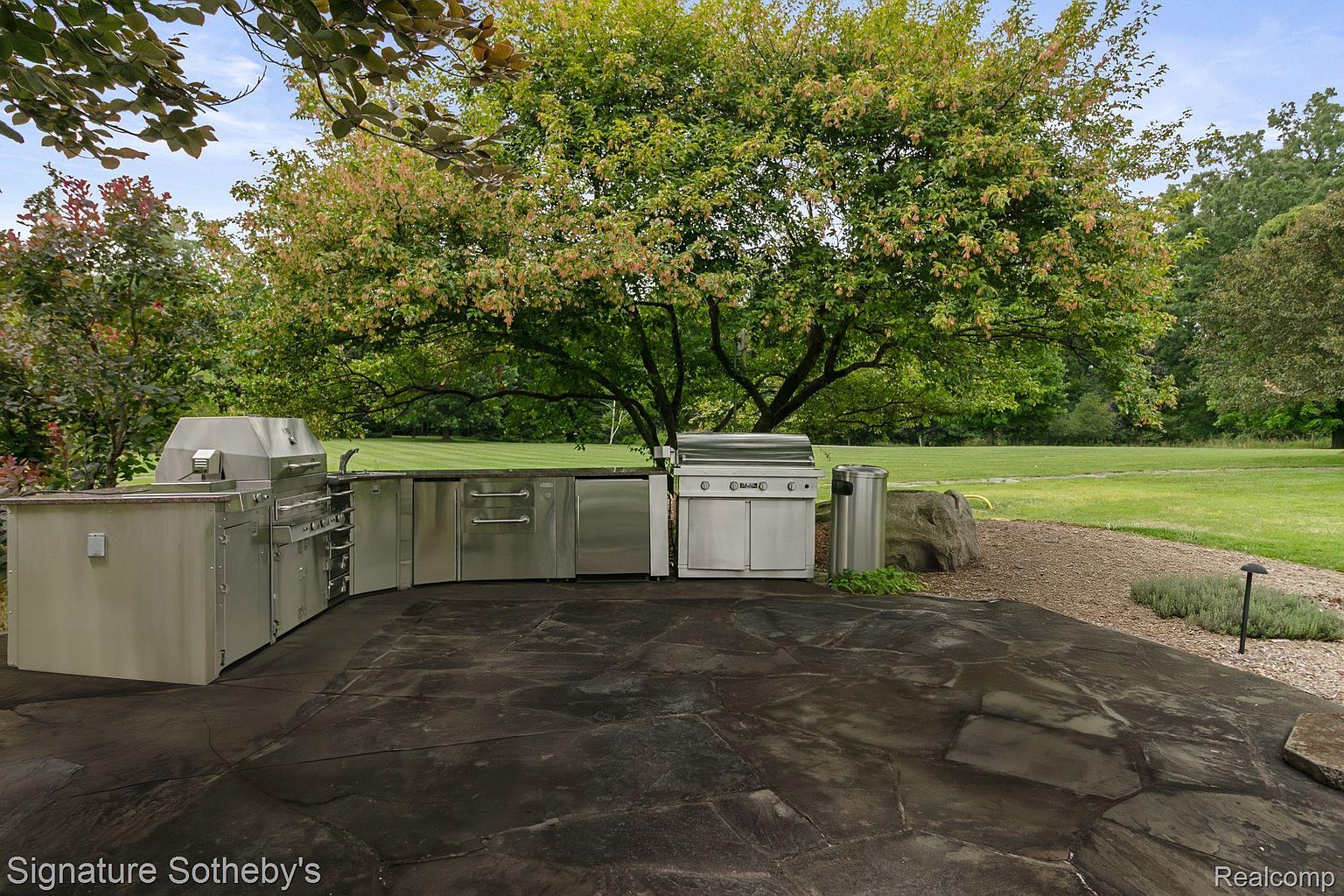 This image showcases an outdoor kitchen area, featuring stainless steel appliances and cabinetry built into a curved layout. The patio is paved with dark, irregular stones, and a large tree provides shade. The scene overlooks a well-maintained lawn, suggesting a spacious and private backyard.