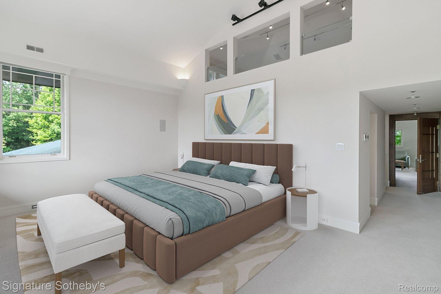 This is a primary bedroom featuring a large bed with a brown upholstered headboard and a teal and gray color scheme. The room has high ceilings with clerestory windows, an abstract art piece above the bed, and a white bench at the foot of the bed. The overall impression is modern and serene, with ample natural light.