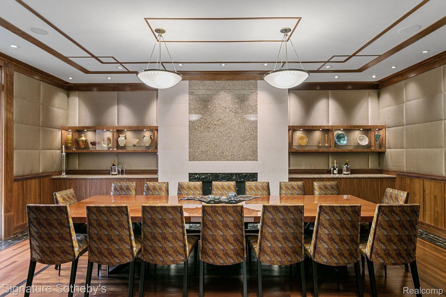 This is an interior shot of a formal dining room. The room features a long wooden table surrounded by upholstered chairs, two pendant lights hanging above the table, and built-in shelving units displaying decorative items. A stone accent wall with a fireplace adds a focal point to the room, creating an elegant and sophisticated atmosphere.