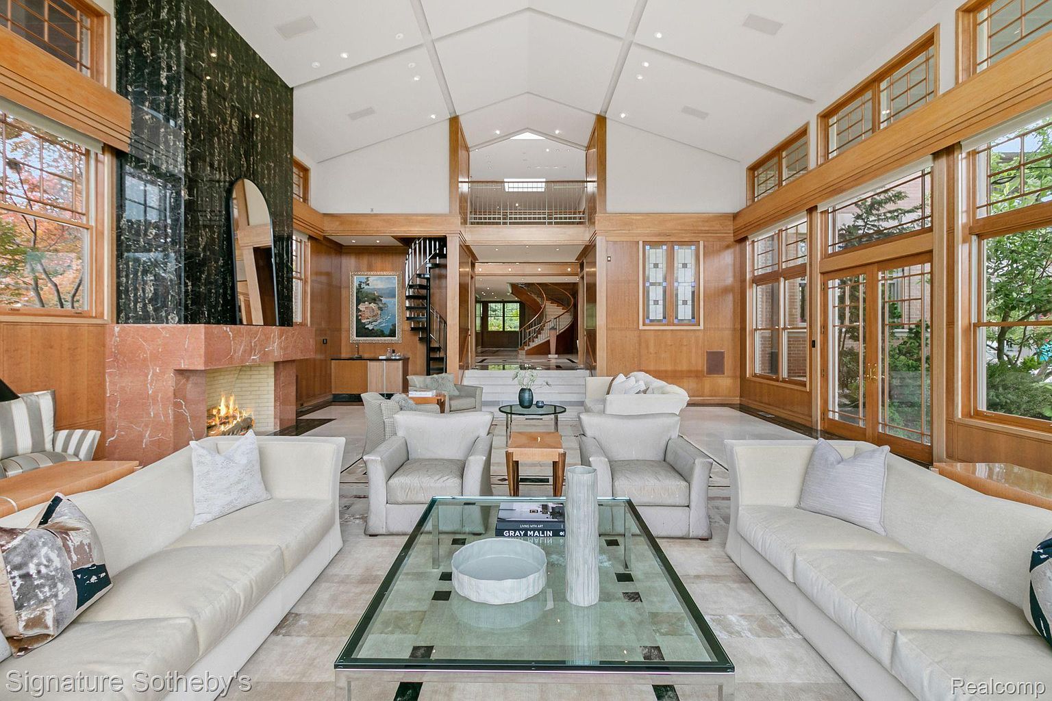 This grand living room features soaring ceilings, wood paneling, and a striking fireplace with a dark marble accent. The space is furnished with multiple white sofas and armchairs, arranged around a large glass coffee table. A spiral staircase and a view into another room with a curved staircase add to the luxurious feel of the home.