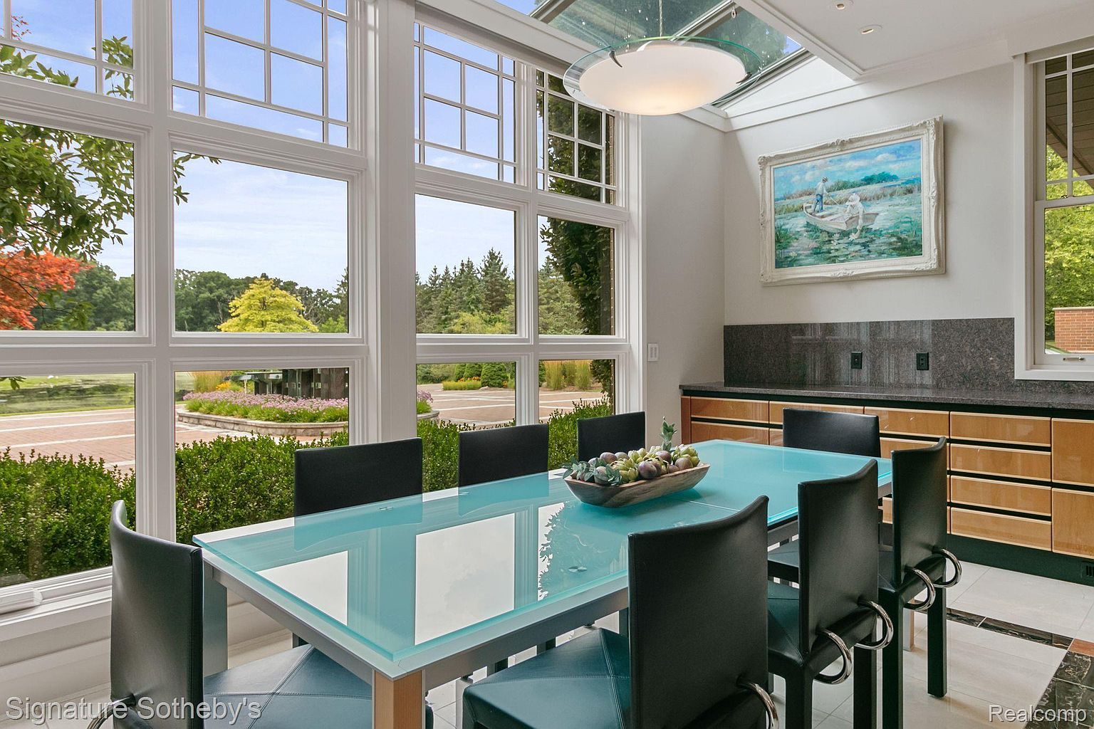 This is an interior shot of a dining room featuring a modern glass-top table with black leather chairs. Large windows offer a view of the landscaped exterior, and a built-in cabinet with a granite countertop adds functionality. A contemporary light fixture hangs from the ceiling, and artwork adorns the wall, creating an elegant and inviting atmosphere.