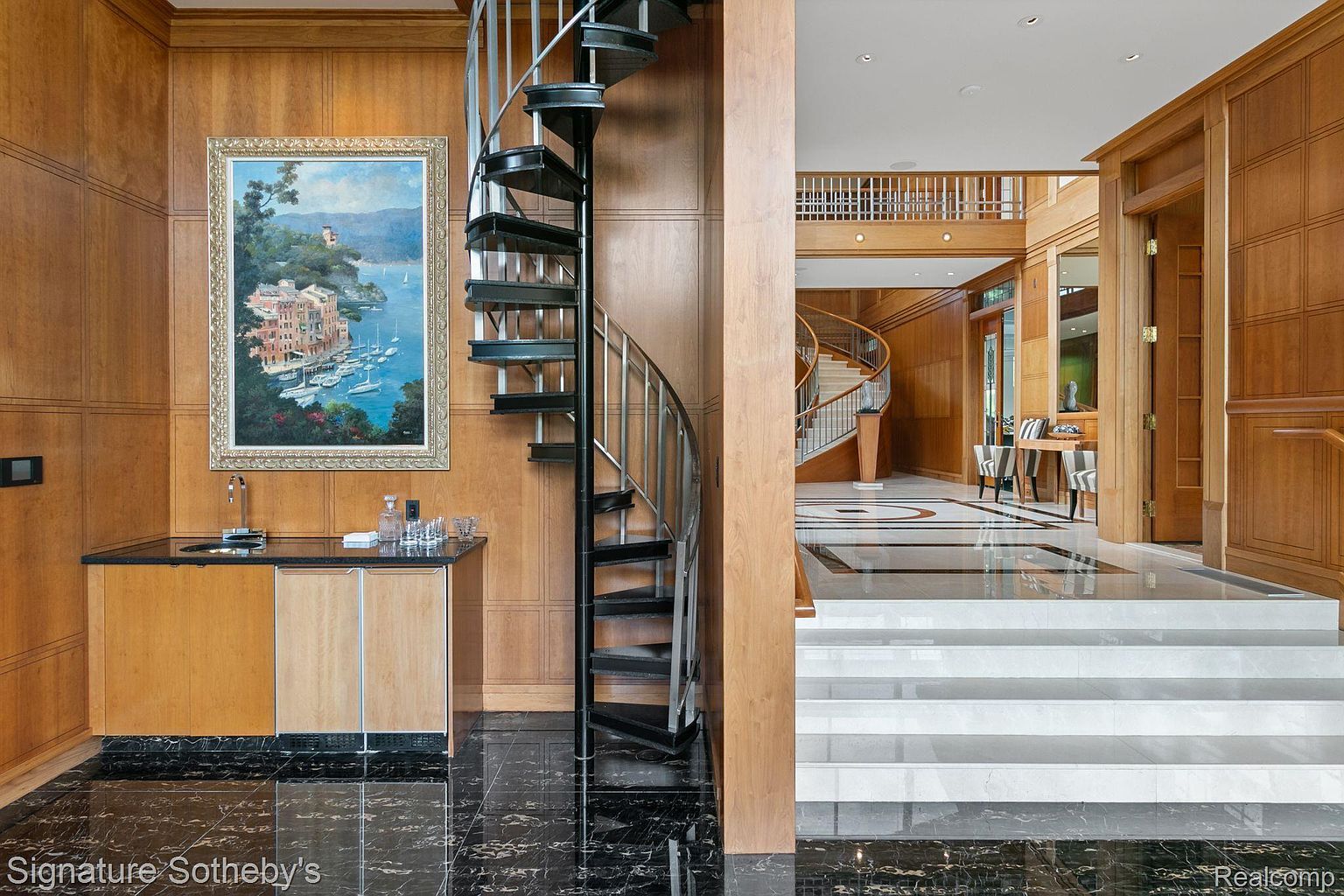This interior shot showcases a grand hallway featuring a spiral staircase and a traditional staircase in the background. The hallway is adorned with wood paneling, a built-in bar area, and elegant marble flooring. The overall impression is one of luxury and sophisticated design.
