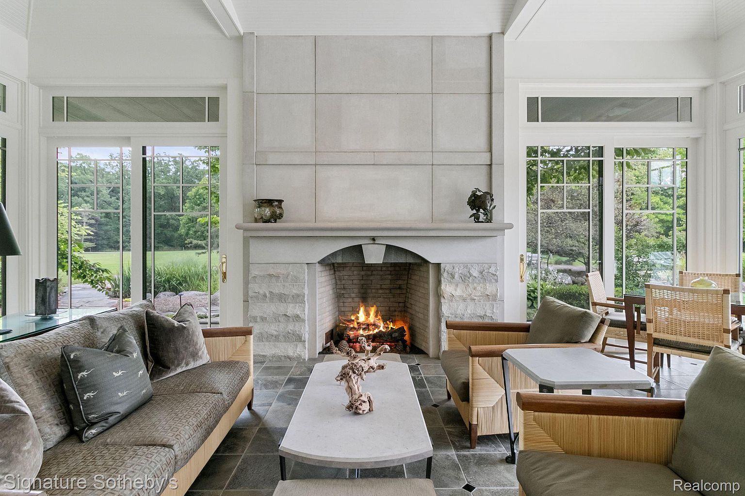 This is an interior shot of a sun-drenched living room featuring a grand stone fireplace as its focal point. The room is furnished with comfortable seating, including a sofa and armchairs, arranged around a unique coffee table. Large windows and glass doors offer views of the lush outdoor landscape, creating a seamless connection between indoor and outdoor living.