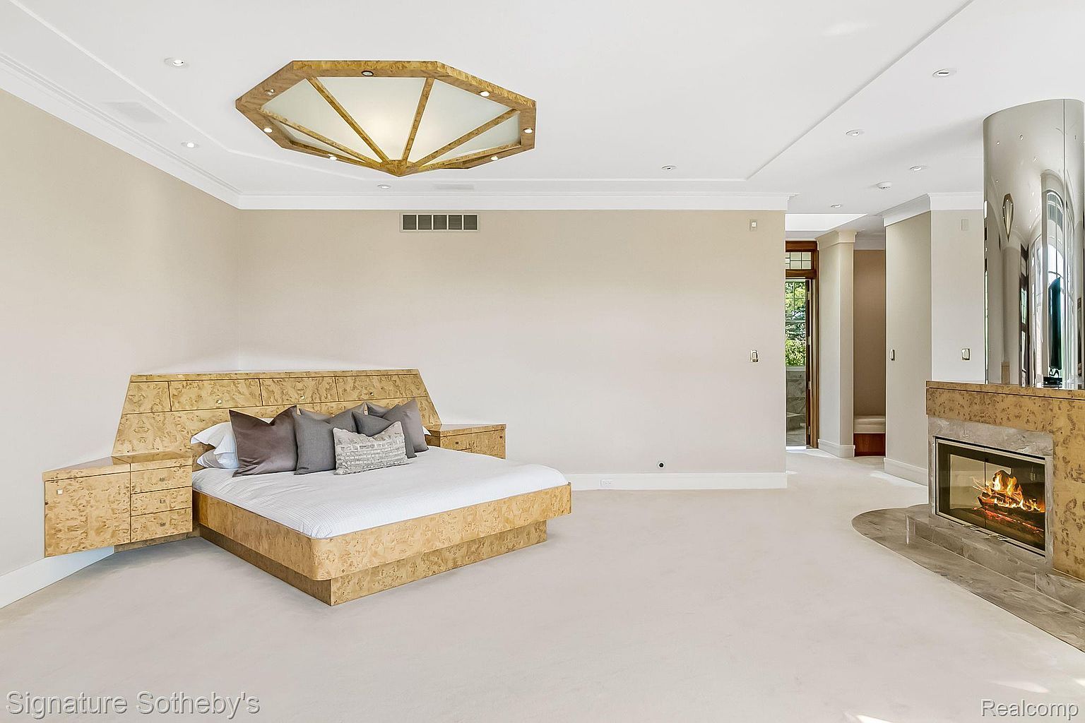 This is a spacious primary bedroom featuring a unique, built-in bed frame and headboard with integrated side tables. The room has neutral-toned walls, light carpeting, and a modern fireplace with a stainless steel chimney. A large, octagonal ceiling light fixture adds a focal point to the room.