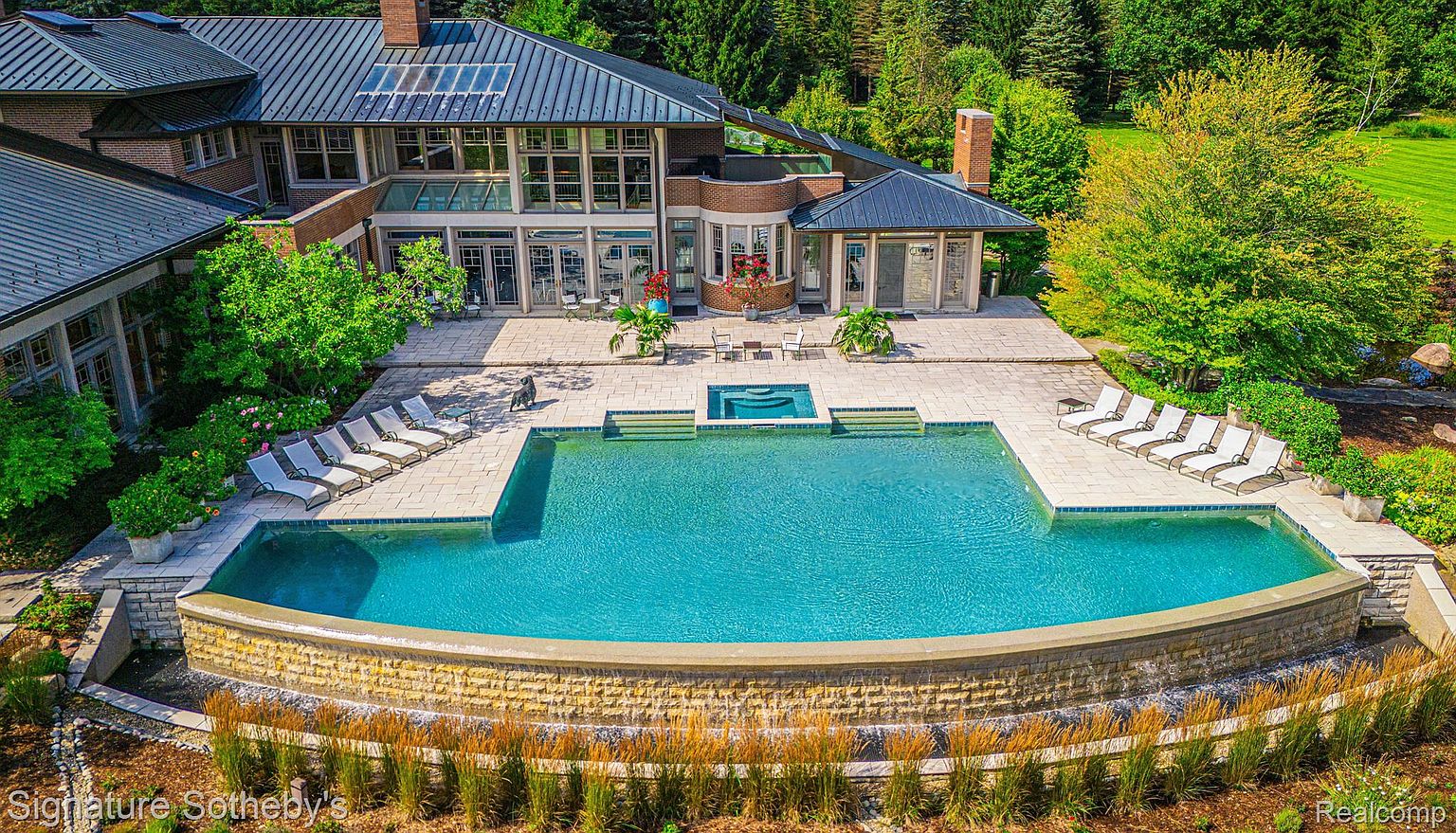 This image showcases a luxurious backyard featuring a large, curved swimming pool with a stone waterfall feature. The pool is surrounded by a spacious patio area with lounge chairs, and a spa is integrated into the design. The house, visible in the background, has large windows and a modern architectural style, complemented by lush landscaping and mature trees.