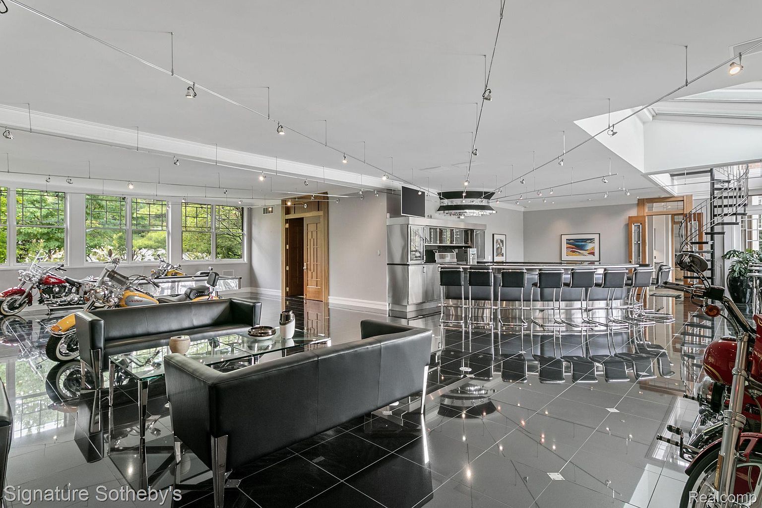 This expansive living room features a sleek, modern design with a black tile floor and track lighting. The room includes a bar area with stools, a seating area with black leather sofas and glass tables, and several motorcycles displayed as art pieces. A spiral staircase is visible in the background, adding to the unique and luxurious feel of the space.
