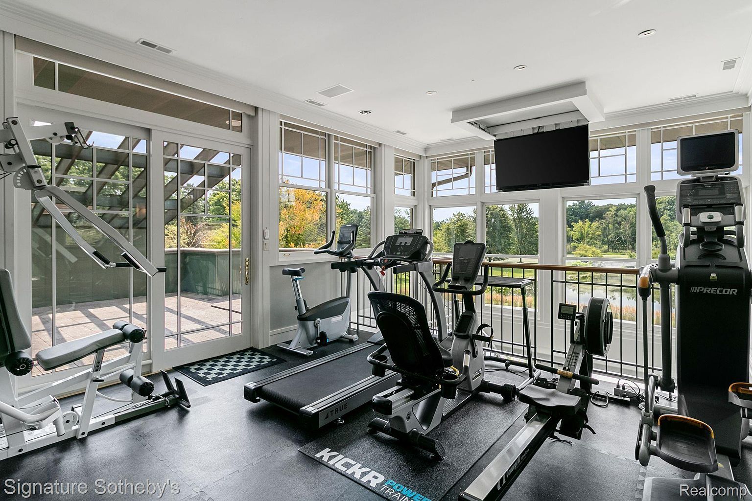 This is an interior shot of a well-equipped home gym. The room features various exercise machines, including a treadmill, elliptical, rowing machine, and weight training equipment. Large windows offer natural light and views of the outdoors, enhancing the workout experience.