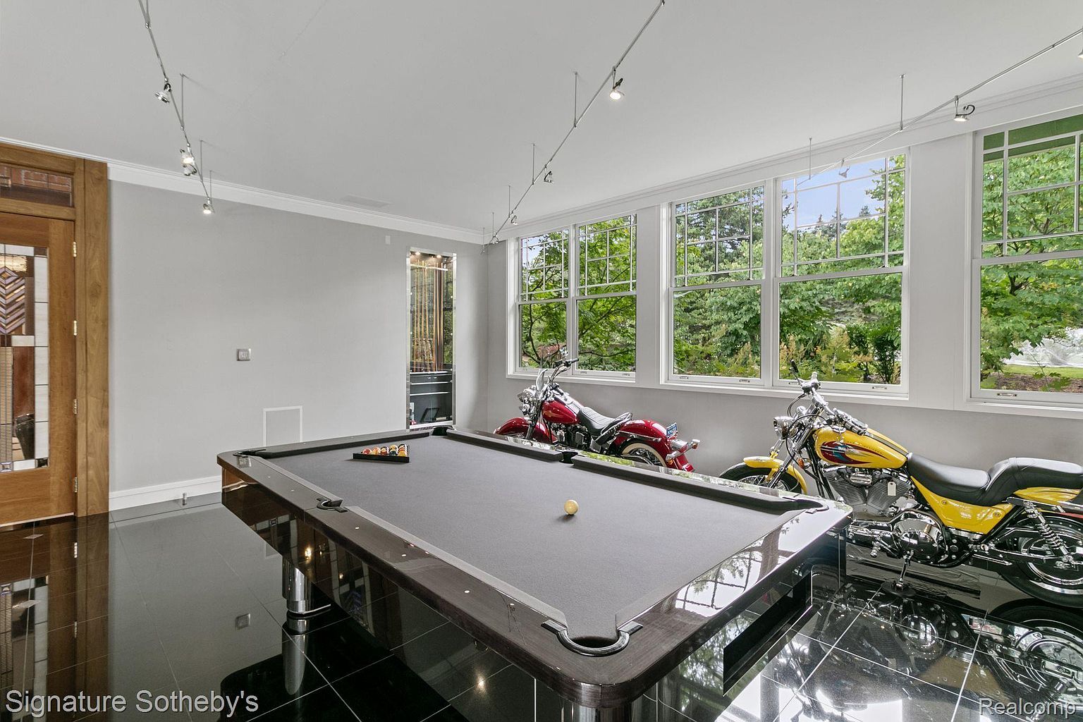 This interior shot showcases a recreational room featuring a pool table as the central element. Two motorcycles are parked near the windows, adding a unique touch to the space. The room is well-lit with natural light and track lighting, and the black tile flooring creates a sleek, modern aesthetic.