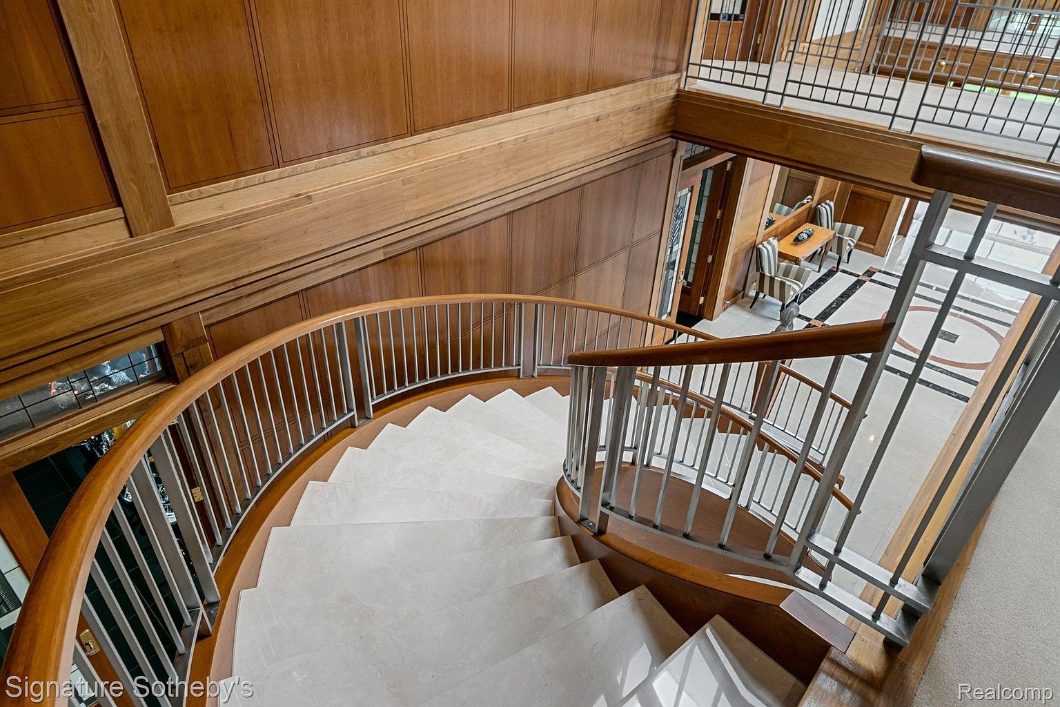 This interior shot showcases a beautifully designed spiral staircase with light-colored steps and a metal railing with wooden accents. The surrounding walls are paneled with rich wood, adding warmth and elegance to the space. The staircase leads to an upper level with a view of a dining area, highlighting the home's architectural details and luxurious finishes.