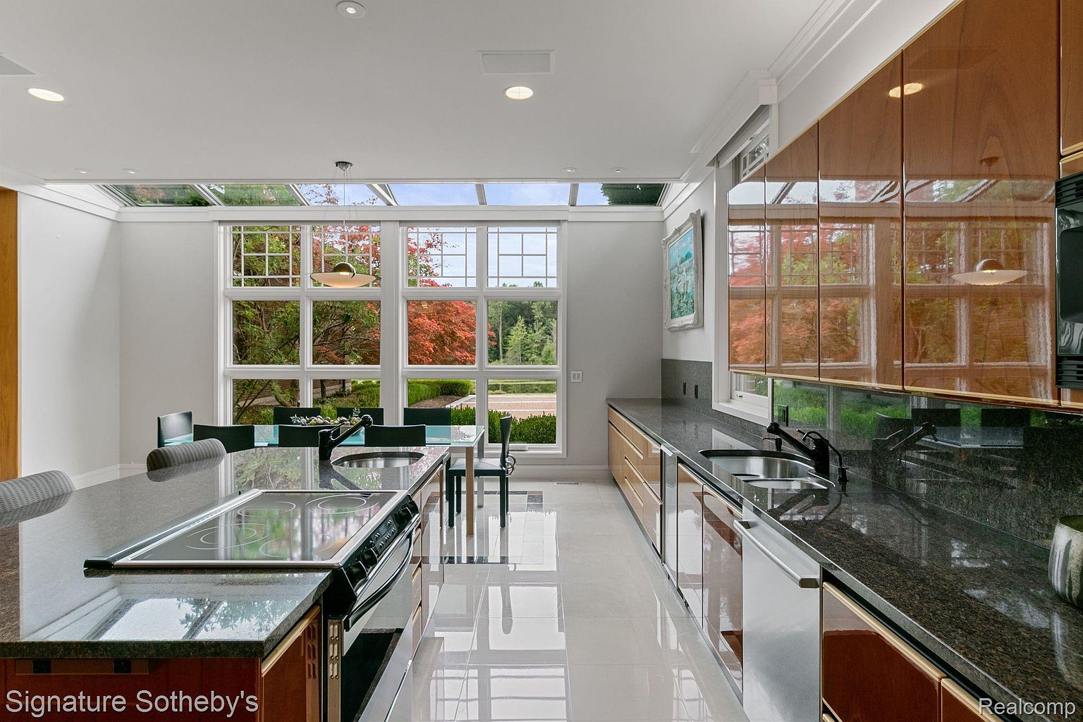 This is a bright and modern kitchen featuring sleek cabinetry, granite countertops, and stainless steel appliances. A large island with a cooktop and sink provides ample workspace, while a wall of windows offers natural light and views of the outdoors. The glossy tile flooring adds to the contemporary aesthetic.