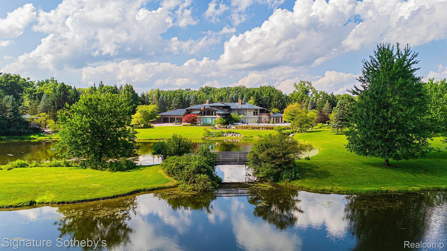 This aerial view showcases a luxurious estate with a sprawling lawn, mature trees, and a serene pond crossed by a bridge. The modern home features a dark roof and multiple wings, suggesting ample living space. The landscape is meticulously maintained, creating a park-like setting that enhances the property's appeal.