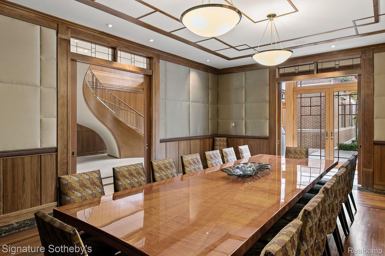 This is an interior shot of a dining room featuring a long, glossy wooden table surrounded by upholstered chairs. The room is decorated with wood paneling, padded wall sections, and unique light fixtures. A curved staircase is visible through an adjacent doorway, adding an elegant touch to the space.