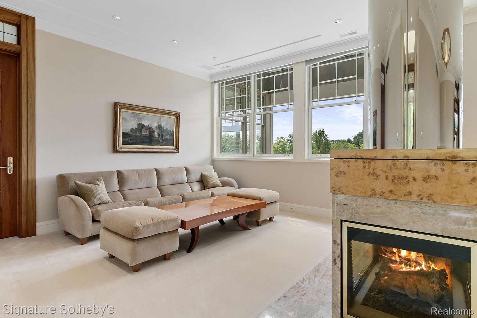 This living room features a comfortable beige sofa with matching ottomans, a wooden coffee table, and a fireplace with a unique marble and wood surround. Large windows provide natural light and a view of the outdoors, while an ornate painting hangs above the sofa. The room exudes a sense of warmth and sophistication.