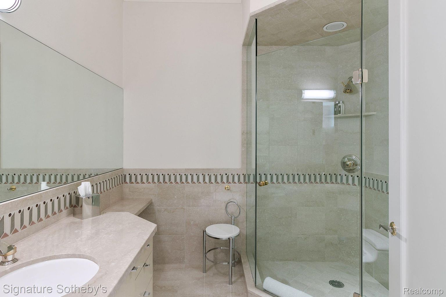 This is a well-lit bathroom featuring a glass-enclosed shower, a vanity with a marble countertop, and a decorative tile border. The shower has a built-in shelf and a chrome showerhead. A small stool is placed near the shower, and a large mirror is above the vanity, creating a bright and functional space.