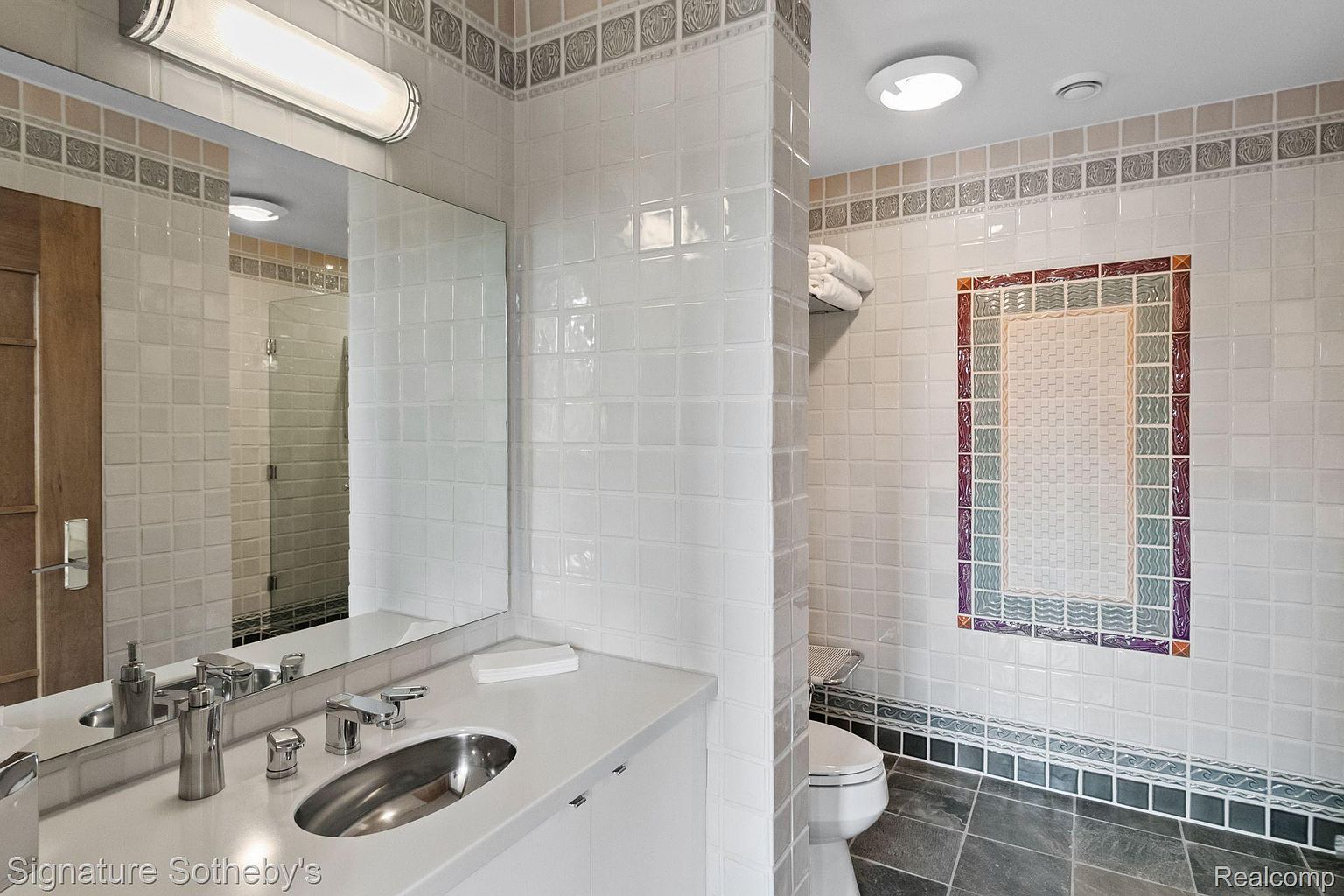 This is a well-lit bathroom featuring white square tiles on the walls, complemented by decorative tile borders near the ceiling and floor. The vanity has a white countertop with a stainless steel sink and modern fixtures. A toilet is visible in the background, and the overall impression is clean and classic.