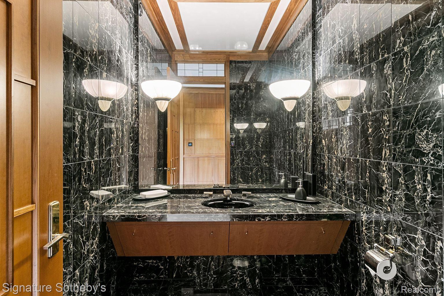 This is a bathroom featuring black marble walls and flooring, creating a luxurious and dramatic atmosphere. A floating wooden vanity with a black countertop and sink is centered in the room, illuminated by sconce lighting on either side of a large mirror. The space is modern and sophisticated, with high-end finishes.