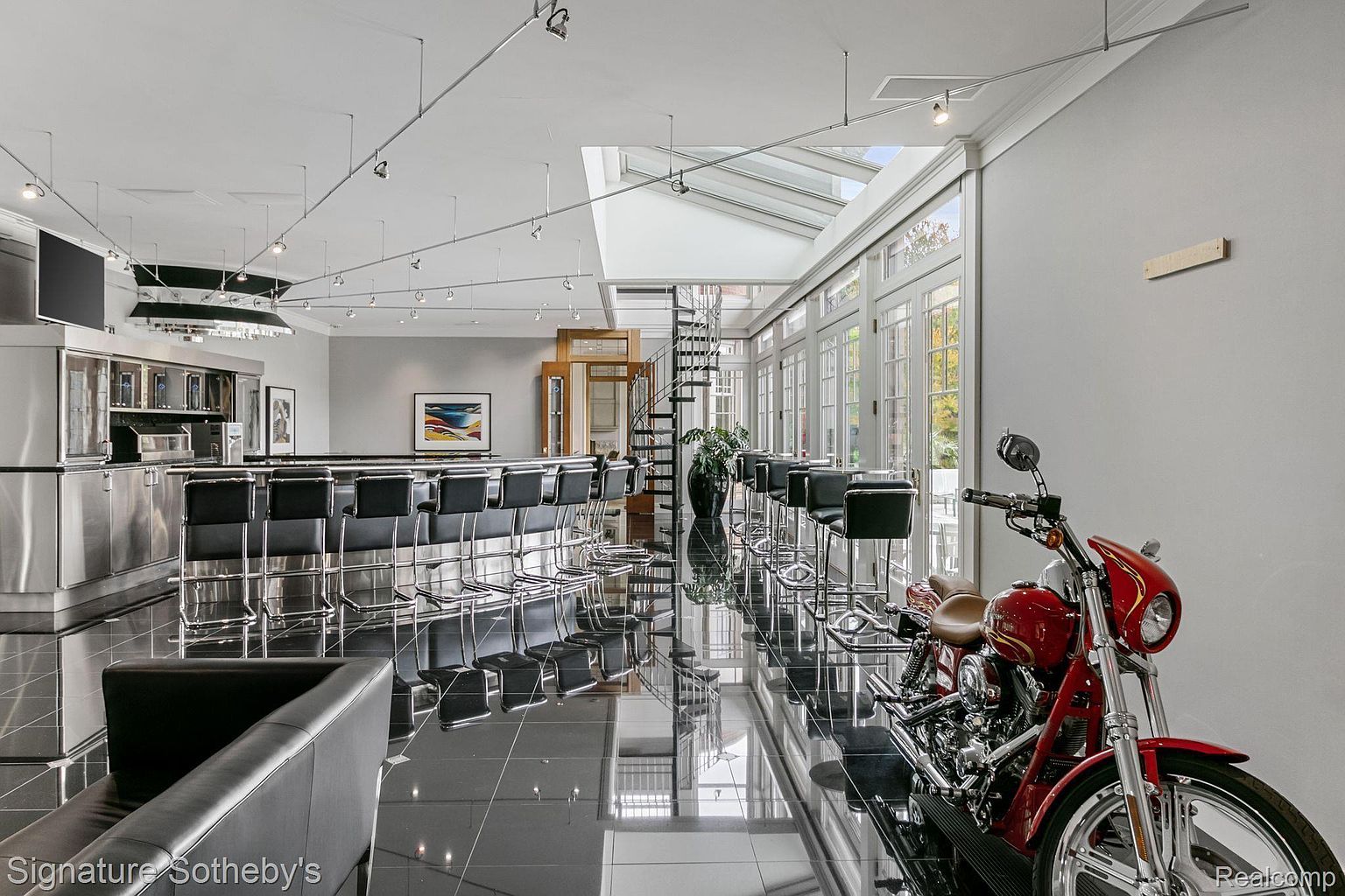 This interior shot showcases a modern living space with a sleek, black tiled floor that reflects the light, enhancing the room's spaciousness. A long bar with black stools lines one side, while a red motorcycle adds a unique, stylish touch. The room is well-lit, featuring a skylight and multiple windows, creating a bright and inviting atmosphere.